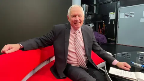 Presenter Nick Owen sitting on a red sofa in a TV studio, wearing a suit and smiling.