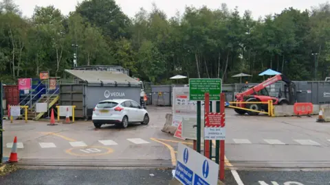 The entrance to a tarmaced areas, with a number of large containers within, as well as a white car and a number of traffic cones, with trees in the background