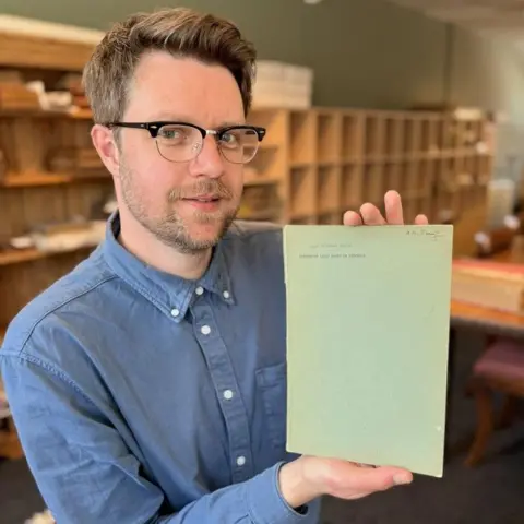 Rare Book Auctions A man with short brown hair, glasses and a blue shirt, carefully holds up an old paper while standing in an archive office.