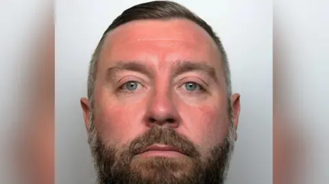 Police mugshot of a man with blue eyes and a short beard