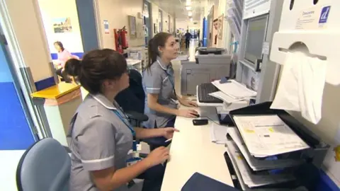 Two nurses with long black hair wearing light blue nurses' uniforms, sit at a desk in a hospital corridor at Northampton General. There is a brown in tray stack in the foreground, there are clipboards on the desk and a photocopier stands in the background. {people are visible a long way down the corridor, and a woman is sitting in a room with blue chairs to the left.