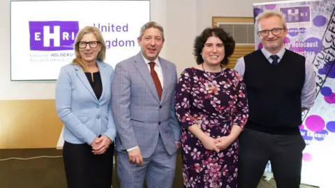 Two women, one in a suit and the other in a flowery dress, and two men in shirts and ties smiling at the camera in front of a banner poster for the EHRI