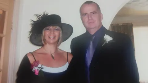 Family Photo Chris and Jane when they first started fostering. Jane is wearing a black top and hat, is smiling at the camera and standing next to Chris, who is wearing a black shirt and blue shirt and tie. They both have flowers pinned to them, as though they are attending a wedding.