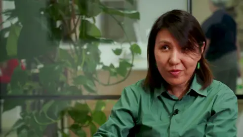 Shamim Saraby in a green shirt speaks during an interview, with office foliage behind her