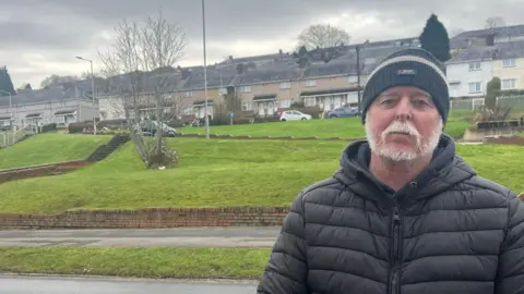 Paul Bennett, who has a white beard and moustache, wearing a black beanie hat and black puffer jacket and stood in front of Gors Avenue