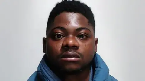 Head-and-shoulders portrait of a person wearing a blue padded jacket, facing the camera against a plain light-colored background. The image is closely cropped around the face and upper shoulders.