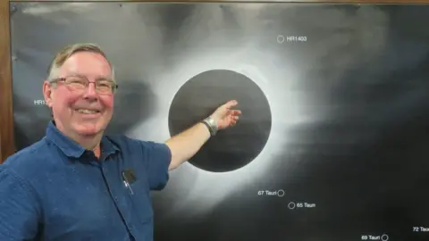 JONATHAN CLOUGH Jonathan Clough pointing at a diagram of a solar eclipse. He is the left of the image, wearing a blue shirt and smiling. 