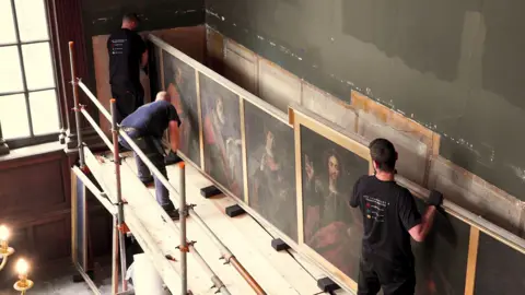 Wentworth Woodhouse Preservation Trust Three men, standing on a scaffolding tower, are lifting a panel containing oil paintings from the wall of the chapel.