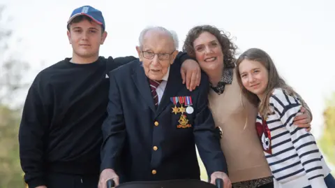 PA Media 99-year-old war veteran Captain Tom Moore, with (left to right) grandson Benji, daughter Hannah Ingram-Moore and granddaughter Georgia, at his home in Marston Moretaine, Bedfordshire, after he achieved his goal of 100 laps of his garden