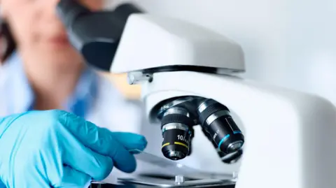 A female scientist using microscope in laboratory. She is wearing a blue shirt and blue plastic safety gloves