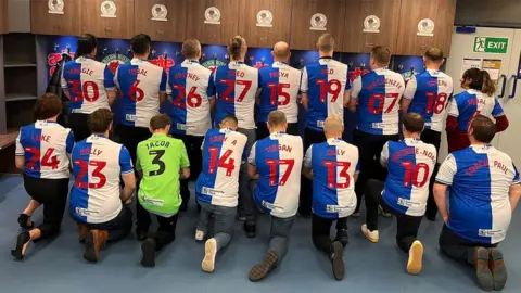 17 members of Dads FC kneeling in two rows in the changing rooms display the backs of their Rovers shirts - half blue, half white - with the names of their children on their shirts