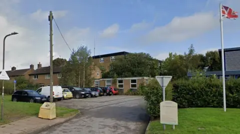 Google Uttoxeter Road (West) car park, Blythe Bridge, where parking charges could be introduced. There is an open space near a single storey building with eight vehicles parked along one side.