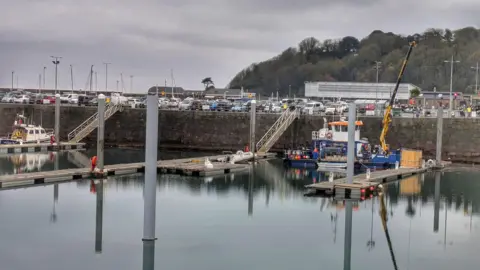 Guernsey Ports A port is seen with numerous vehicles parked on a car park, and a large building behind, with a tree-lined cliff in the background. In the foreground is a body of water, with decking and a crane on a boat. 