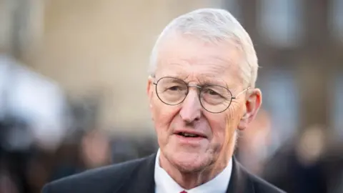 PA Media Hilary Benn is an older man with short white hair. He is wearing a dark suit, white shirt and round metal framed glasses.