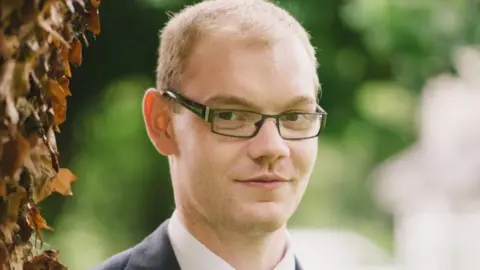 The family's handout Dennis Sheer is wearing a rectangular glasses as he stares on the camera. The background is blurred. Dennis has light thin hair and wearing a white shirt and deep jacket.