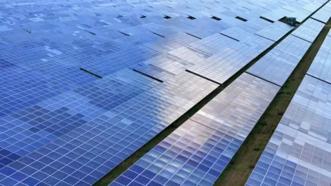 Getty Images Rows of shiny black solar panels are lined up across a green field in a solar farm, each of them reflecting the sunlight