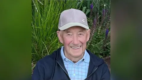 Albert Butterfield Bert Butterfield is smiling at the camera. He is wearing a baseball cap and a blue jacket. He is sitting in front of plants.
