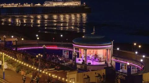 An audience watch musicians perform on the Eastbourne Bandstand on the seafront in Eastbourne, UK, on Friday, Aug. 4, 2023. The bandstand is lit up with purple lights, while the pier in the background is illuminated in white light.