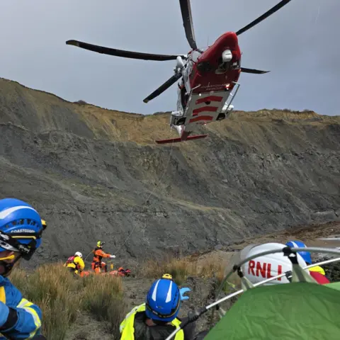 Kimmeridge Coastguard Rescue Team RNLI and coastguard teams at a cove with a helicopter above as they winch down a stretcher.