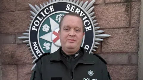 A police officer is talking to the media. Behind him is the PSNI emblem. The PSNI emblem is attached to a brick wall. It’s Saint Patrick’s saltire with six symbols: the scales of justice, a harp, a torch, a laurel leaf, a shamrock, and a crown.