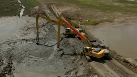 John Fairhall/BBC Two large yellow diggers work on lifting and shaping mud from trucks on a narrow road onto the wetlands next to it 