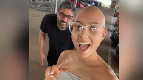 PA Media Asha with her father, Jayesh. They are walking in a multi-storey car park. Asha is wearing glasses and has a shaved head following her treatment. She has a shoulder-less silver dress on and has her mouth open while doing a peace sign with her fingers. Jayesh is crouched in the background smiling while wearing a black T-shirt and glasses.