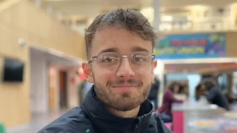 Alex Dunlop/BBC A young man with short brown hair and glasses smiles at the camera in a head and shoulders image. He has a short brown beard and is wearing a black jacket which is zipped up.