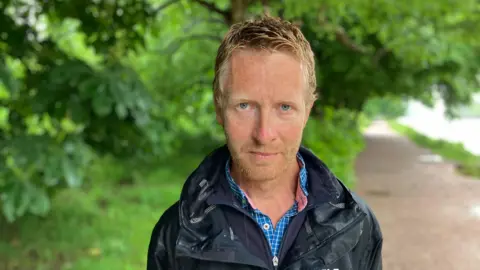A man stands on a riverside pathway, there are trees behind him. He is wearing a black waterproof jacket which is wet from rain. 