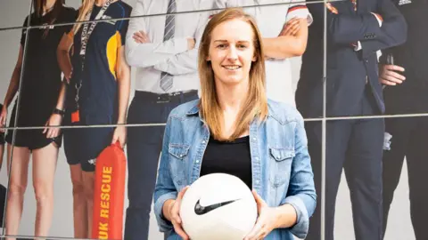 Kerys Harop is standing holding a football in front of a large sports‑themed mural outside the Walsall Sports Centre at the University of Wolverhampton. She has blonde hair and is wearing a denim jacket over a black top