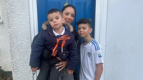 A mother and her two sons look relieved as they stand outside their blue front door. 