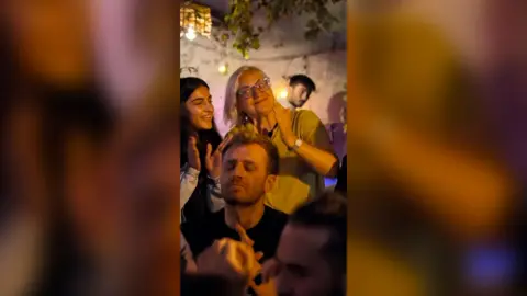 Two women are clapping in an outdoor group setting at night. One is young and the other is older. People dance in front of them.