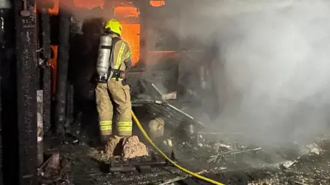 A lone firefighter working to extinguish a fire at an outbuilding. The glow of the flames is illuminating the inside of the building and smoke is visible.