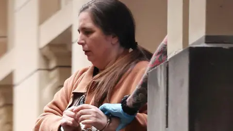 Getty Images A woman in a brown jacket with a brown ponytail with her wrists in handcuffs. Her left wrist is being held by a gloved hand on a tattooed arm emerging from behind a pillar
