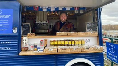 Mike Lowe is smiling at the camera. He was dark hair and beard and is wearing a checked shirt, at the inside counter of a blue mobile coffee stall. Two shelves in front of him on the customer side contain flat basket trays and a row of jarred items. To the right, a blue sign says San Seb coffee van parking only.