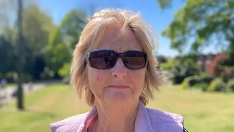 A head and shoulders photograph of Pauline Gray who is wearing a blue and white top and a lilac coloured gilet. She is standing in a park.