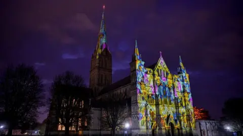 Finbarr Webster The front of a grand cathedral at night covered in a coloured light projection, the colours are yellow, blue and purple