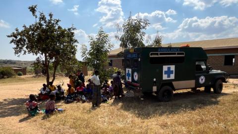 People wait outside the ambulance under the shade of a tree for treatment in rural outreach clinics