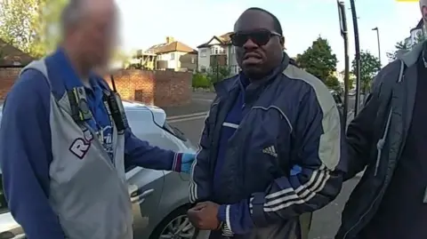 Metropolitan Police Bodycam footage showing Simon Levy in a navy blue Adidas jacket and sunglasses being detained by police officers on a suburban street. An officer on the left, who's face is blurred, wearing a blue shirt and grey jacket, fitted with a body-worn camera and radio, holds the man's arm.