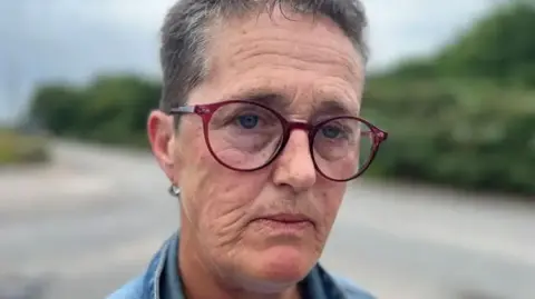 Close up of Allison Rowe who has short greying hair wearing maroon rimmed round glasses and a denim top. She is photographed in Fleetwood.