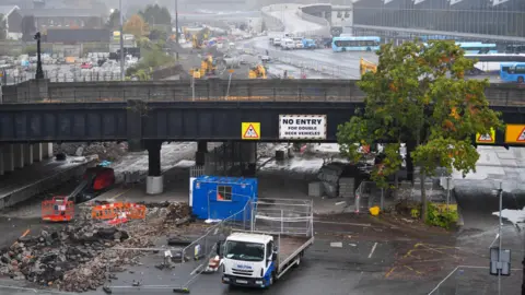 Pacemaker Dismantling of Boyne Bridge. There is a work van parked next to fencing. Bricks have been dug up from the ground. Grand Central Station can be seen in the background. The ground is wet.