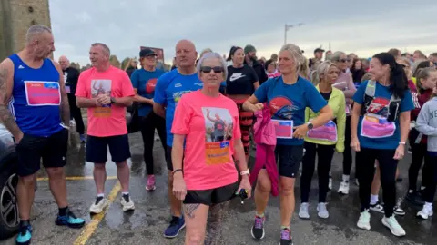 Dozens of people in pink and blue running gear are chatting as they wait for the run to begin. Standing at the forefront of the picture, Karen McNeill Smith is wearing a pink running top with a picture of Sam on it. She has short grey/ purple hair.