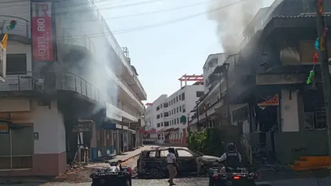 City of Puerto Vallarta A burnt car blocking a road where smoke can be seen rising from a hotel and convenience store