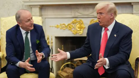 Micheál Martin sits in the Oval Office in a chair opposite Donald Trump. Martin is speaking and has his arms open. Trump is sat with his hands held and is looking at Martin.