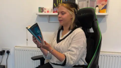 A woman sitting in a computer chair reading a book. She has shoulder-length hair that has a blonde streak. She also has a nose ring and is wearing a white V-neck jumper with a black V.