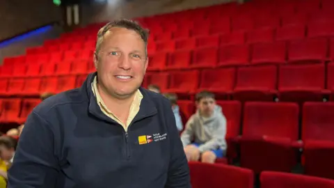 Philip smiles at the camera as he wears a blue quarter zip jacket with a yellow shirt underneath. He has short black hair and behind him are red theatre chairs and some children are sitting on them.