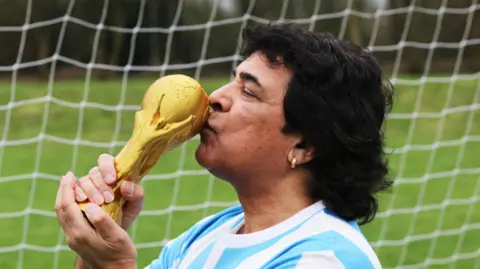 A man resembling Diego Maradona is stood in front of a football goal kissing a replica of the golden World Cup trophy. He is dressed in a blue and white football shirt, has thick black hair and wears an earing.
