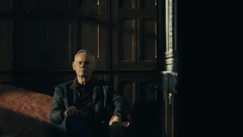 5 Broadcasting Limited / Paramount / Wonderhood Studios/ PA Clunes sat on a red sofa. He wears a brown jumper and black coat