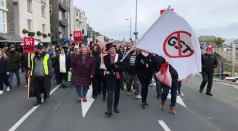 BBC A crowd walking towards the camera as a man in a hat brandishes a flag with a the letters GST and a red strikethrough. 