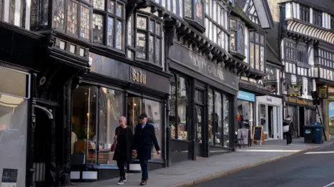 A street running up a hill with a series of black and white timbered buildings with shop windows running up the left hand side and a man and a woman walking down the hill