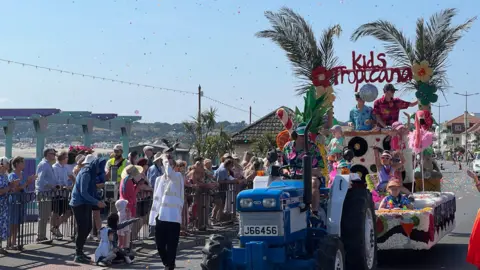 A decorated blue tractor pulls a colourful parade float along a seaside road as crowds watch from behind barriers. The float is decorated with tropical-themed flowers and palm leaves and displays the sign “Kids Tropicana.” Confetti fills the sky above the parade route on a bright sunny day.